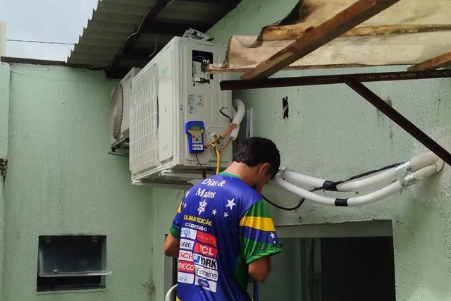 Técnico uniformizado da Dias & Matos sobre escada, conectando a unidade externa de um ar-condicionado split instalada na parede de uma residência
