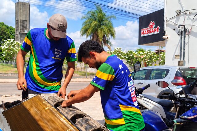 Dois técnicos uniformizados realizando a limpeza de um ar-condicionado split piso-teto desmontado em frente a um estabelecimento comercial